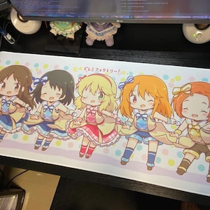 May include: A rectangular mousepad featuring five anime-style characters in colorful outfits. The characters are holding hands and smiling, set against a pastel background with the text "ドレミファクトリー!".