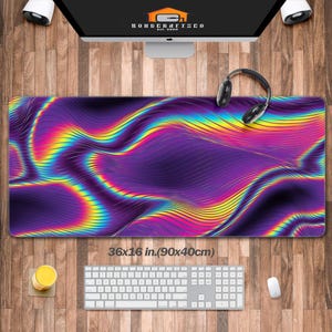 Puede incluir: Una alfombrilla de escritorio rectangular grande con un diseño ondulado vibrante en tonos morado, amarillo y verde. La alfombrilla mide 90 x 40 cm. Un teclado, un ratón y unos auriculares están sobre el escritorio de madera.