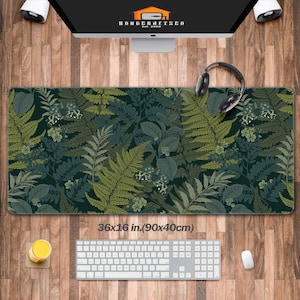 Puede incluir: Alfombrilla de escritorio rectangular con un diseño botánico de helechos y hojas en tonos verdes y verde azulado. La alfombrilla mide 90 x 40 cm. Un teclado, un ratón y un vaso de líquido amarillo están sobre el escritorio de madera.