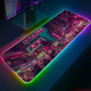 Puede incluir: Un ratón de ordenador negro con un borde LED arcoíris, impreso con una escena de ciudad de neón con luces rosas y azules.