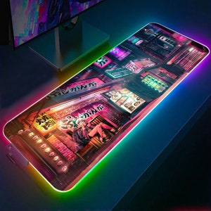 May include: A colourful mouse mat with a neon glow around the edges. The design features a Japanese cityscape with neon signs and a character sitting on a ledge. The text on the signs reads "おつかれさま".