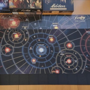 May include: Firefly board game components, including game boards and expansion sets, are displayed on a wooden surface. The game boards feature a star map design with various celestial bodies and the text "Firefly The Game".