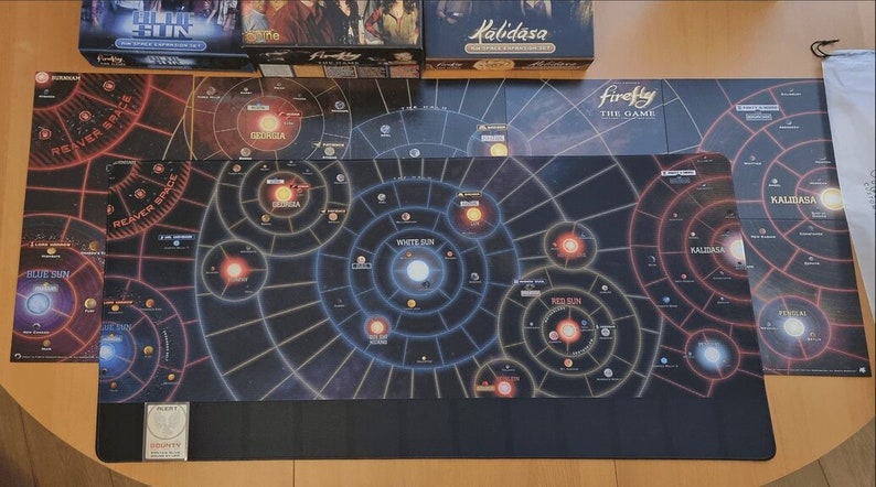 May include: Firefly board game components, including game boards and expansion sets, are displayed on a wooden surface. The game boards feature a star map design with various celestial bodies and the text "Firefly The Game".