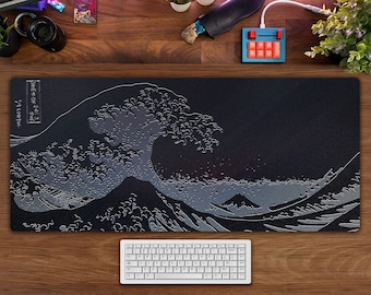 May include: A black mousepad with a white graphic of the Great Wave off Kanagawa, a famous Japanese woodblock print by Hokusai. The mousepad is on a wooden desk with a white keyboard and a red and blue keypad.