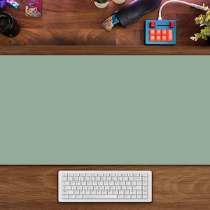 Puede incluir: Una alfombrilla de escritorio verde con un teclado blanco sobre un escritorio de madera. La alfombrilla de escritorio es de color verde claro y el teclado es un teclado blanco estándar con teclas negras.