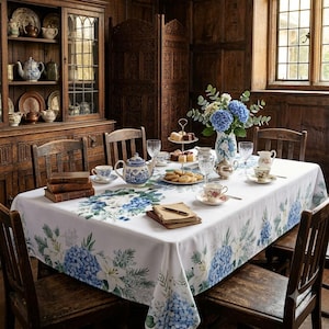 May include: A white rectangular tablecloth featuring a blue and green floral print of hydrangeas and lilies. The tablecloth is draped over a dining table set with a blue and white teapot, teacups, saucers, a tiered dessert stand, and a vase.