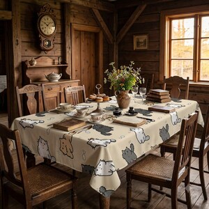 May include: A rectangular tablecloth with a cream background featuring a repeating pattern of cartoon cats in white, gray, and orange. The tablecloth is draped over a wooden dining table surrounded by wooden chairs in a rustic room with wood walls.
