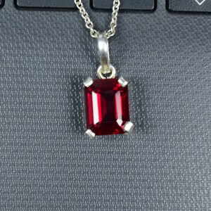 May include: A silver pendant necklace featuring a rectangular, deep red gemstone. The gemstone is set in a silver bezel with four prongs. The chain is a delicate silver link. The pendant is displayed against a dark gray background.