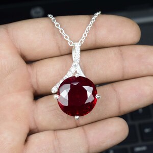 Op de afbeelding: Een zilveren ketting met een grote, ronde, dieprode edelsteen hanger. De hanger is gezet in zilver met kleine, heldere accentstenen. De ketting is een delicate zilveren schakel.