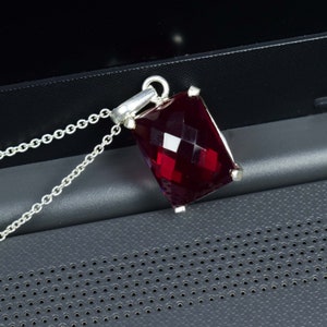 May include: A silver necklace featuring a rectangular, faceted, deep red gemstone pendant. The pendant is suspended from a delicate silver chain. The gemstone has a brilliant cut, reflecting light. The necklace is displayed against a dark background.