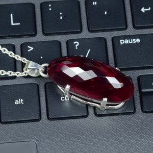 Op de afbeelding: Een zilveren ketting met een grote, facetgeslepen, ovaalvormige, dieprode edelsteen hanger. De hanger is gezet in een zilveren montuur en hangt aan een fijne zilveren ketting. De ketting ligt op een zwart toetsenbord.