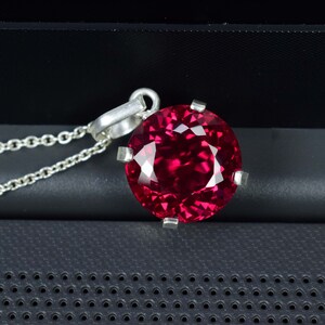 Könnte beinhalten: Eine silberne Anhängerkette mit einem großen, runden, facettierten, tiefroten Edelstein. Der Edelstein wird von vier silbernen Krappen gehalten. Die Halskette ist ebenfalls silberfarben und filigran.