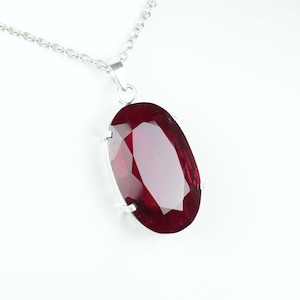 Peut inclure: Un collier argenté avec un grand pendentif ovale en pierre précieuse rouge foncé facettée. Le pendentif est serti dans une lunette argentée et est suspendu à une fine chaîne argentée. La pierre précieuse a une surface brillante et réfléchissante.
