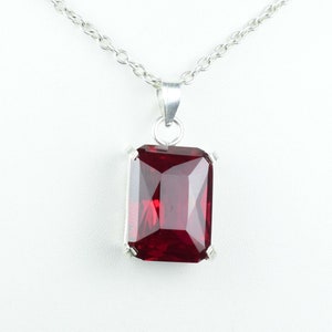 May include: A silver necklace with a rectangular, faceted, deep red gemstone pendant. The pendant is set in a silver frame and hangs from a delicate silver chain. The gemstone has a brilliant cut, reflecting light.