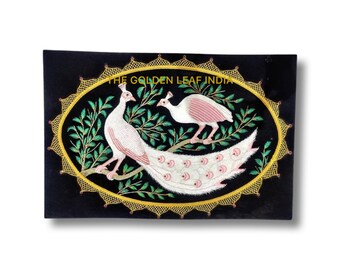 Puede incluir: Obra bordada sobre terciopelo negro con dos pavos reales blancos con detalles en rosa, uno con las plumas de la cola extendidas. La obra está enmarcada con un óvalo dorado y un borde decorativo. El texto "THE GOLDEN LEAF INDIA" es visible en la parte superior.