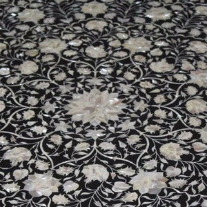 May include: Black surface inlaid with a floral pattern in white mother-of-pearl. The design features stylized flowers and vines, creating a symmetrical, ornate pattern. The overall effect is elegant and decorative.