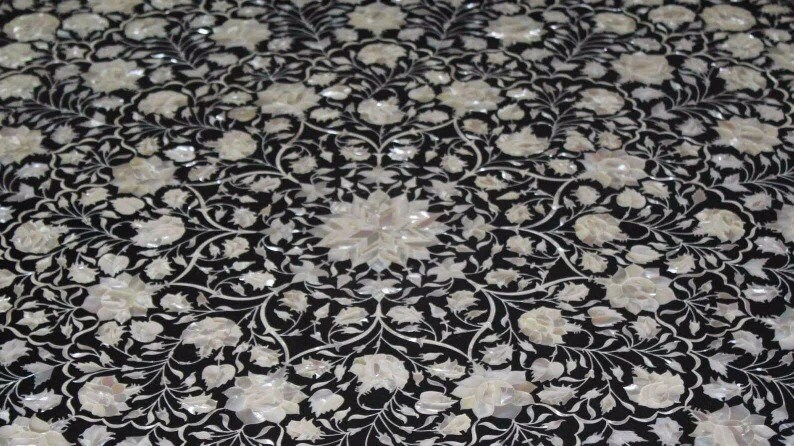 May include: Black surface inlaid with a floral pattern in white mother-of-pearl. The design features stylized flowers and vines, creating a symmetrical, ornate pattern. The overall effect is elegant and decorative.