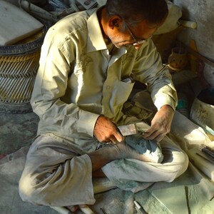 May include: A man sits cross-legged on a floor, using a tool to carve a green stone. He is wearing a light brown shirt and pants. There are several tools and pieces of green stone on the floor around him.