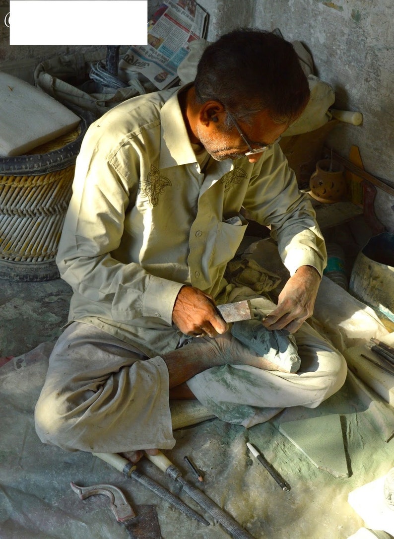 May include: A man sits cross-legged on a floor, using a tool to carve a green stone. He is wearing a light brown shirt and pants. There are several tools and pieces of green stone on the floor around him.