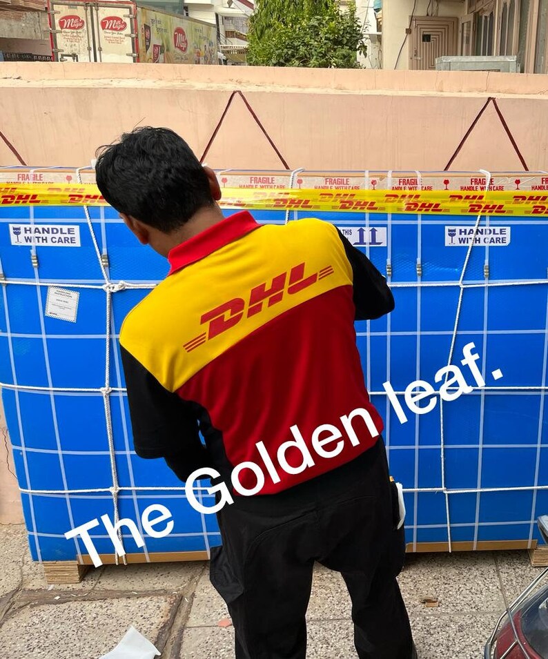 May include: A person wearing a DHL uniform stands near a large blue crate secured with netting. The crate is labeled with "Fragile" and "Handle with Care" warnings. The DHL uniform is yellow, red, and black.