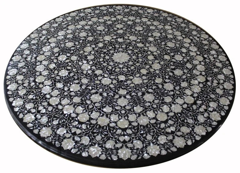 May include: Round black table top with an intricate floral inlay design. The inlay features white and off-white flowers and foliage, creating a striking contrast against the dark background. The table top is approximately 48 inches in diameter.