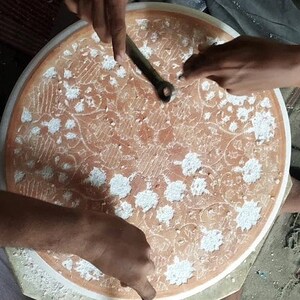 May include: A person is using a tool to carve a circular design into a round, white surface. The surface has a brown background with a floral pattern. The person is wearing a white shirt and has their hands on the surface.