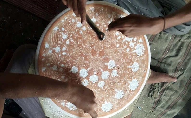 May include: A person is using a tool to carve a circular design into a round, white surface. The surface has a brown background with a floral pattern. The person is wearing a white shirt and has their hands on the surface.