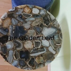 May include: A round table top made of agate stone. The table top is a mosaic of different sized and shaped pieces of agate in shades of grey, brown, and white. The text "The Golden Leaf" is visible on the table top.