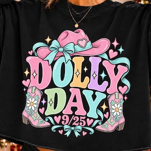 May include: A black long-sleeve cropped sweatshirt featuring a graphic print with the text DOLLY DAY 9/25. The design includes a pink cowboy hat with a bow, two pink cowboy boots with floral accents, and various colorful hearts and stars.