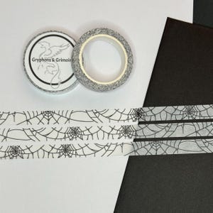 May include: Two rolls of decorative washi tape. One roll is grey with a white pattern, and the other is a grey and white spiderweb design. The tape is labelled "Gryphons & Grimoire". Strips of spiderweb tape are also visible.