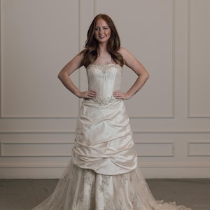 May include: A strapless ivory wedding gown with a beaded bodice and a draped skirt. The skirt flows into a lace-trimmed train. The dress is designed with a fitted silhouette and is suitable for a wedding or formal event.