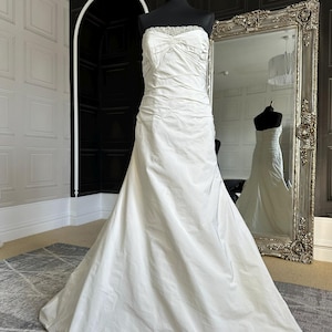 May include: A white strapless wedding dress with a beaded bodice and a flowing skirt. The dress is on a mannequin in a room with a mirror.