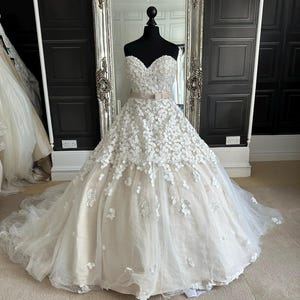 May include: A strapless ivory wedding gown with a sweetheart neckline and a full skirt. The bodice and skirt are adorned with floral appliques. A satin ribbon belt with a bow accents the waist. The dress is displayed on a black mannequin.