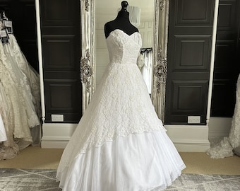 May include: A white strapless wedding dress with a lace bodice and a tulle skirt. The dress is on a mannequin in a room with a mirror and black walls.