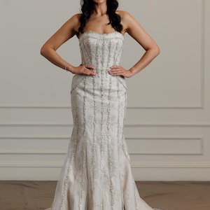 May include: A strapless ivory wedding gown with intricate beaded detailing. The dress features a fitted bodice and a flared mermaid skirt. The gown is floor-length and has a subtle train. The bride is smiling with her arms on her hips.