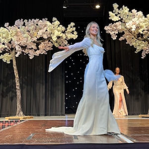 May include: A woman in a white gown with long sleeves and a flowing skirt walks on a stage with a wooden floor. The stage is decorated with two white flowering trees.
