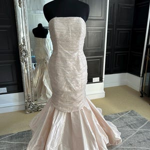 May include: A strapless, blush-colored wedding dress with a fitted bodice and a flowing, layered skirt. The dress is made of a soft, shimmering fabric and features delicate beading.