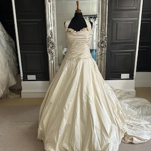 May include: Ivory-colored wedding dress with a halter neckline and a full skirt. The bodice is ruched, and the skirt flows into a train. The dress is displayed on a mannequin in front of a large ornate mirror.