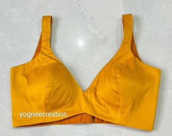 May include: A vibrant mustard yellow blouse featuring a plunging V-neckline and broad straps. The fabric has a slight sheen. The text "yogneecreation" is visible on the lower left. The garment is tailored with a fitted silhouette.