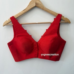 May include: A red sleeveless blouse with a deep V-neckline and a scalloped edge. The blouse is hanging on a wooden hanger. The text "yogneecreation" is printed on the front of the blouse.