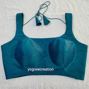 May include: A teal blue sleeveless blouse with a square neckline and a small, decorative tassel on the back. The blouse has a simple design and is made of a smooth fabric.