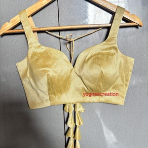Könnte beinhalten: Ein ärmelloses, bauchfreies Oberteil in Gold mit tiefem V-Ausschnitt und einem Quasten-Detail am Saum. Das Oberteil befindet sich auf einem Holzbügel. Der Text "yogneecreation" ist auf dem Oberteil sichtbar.