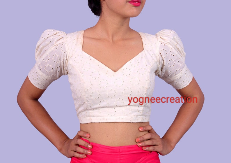 Puede incluir: Blusa color crema con mangas cortas abullonadas y escote en V. La tela presenta un delicado dise&ntilde;o de ojal. La blusa es de estilo corto, y el texto "yogneecreation" es visible en la parte delantera.