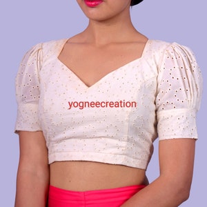 Puede incluir: Blusa color crema con escote cuadrado y mangas cortas abullonadas. La tela presenta un dise&ntilde;o de ojal. El texto "yogneecreation" est&aacute; impreso en la blusa.