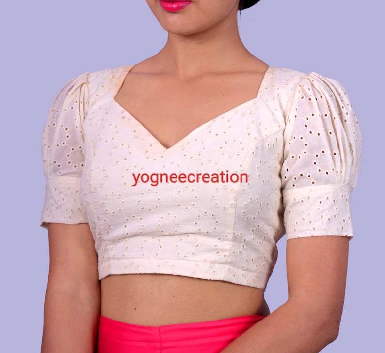 Puede incluir: Blusa color crema con escote cuadrado y mangas cortas abullonadas. La tela presenta un dise&ntilde;o de ojal. El texto "yogneecreation" est&aacute; impreso en la blusa.