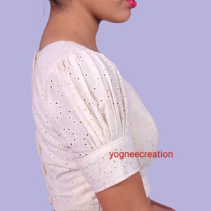 Puede incluir: Blusa calada de color crema con mangas cortas abullonadas y cuello cuadrado. La blusa tiene cierre de botones en la espalda y est&aacute; hecha de una tela ligera. El texto "yogneecreation" es visible en la manga.