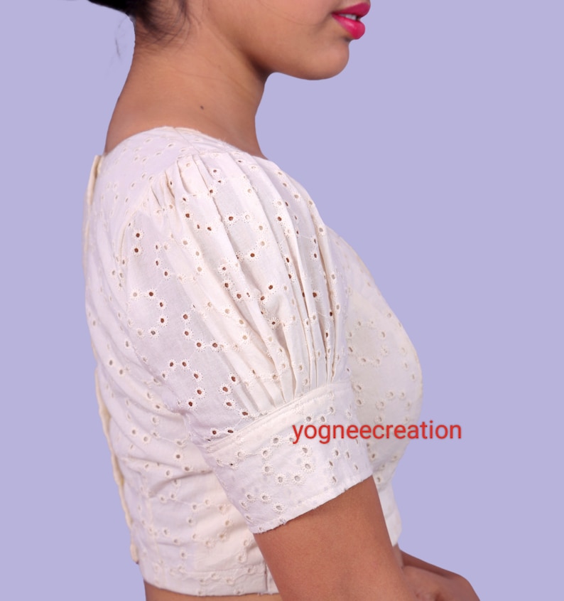 Puede incluir: Blusa calada de color crema con mangas cortas abullonadas y cuello cuadrado. La blusa tiene cierre de botones en la espalda y est&aacute; hecha de una tela ligera. El texto "yogneecreation" es visible en la manga.