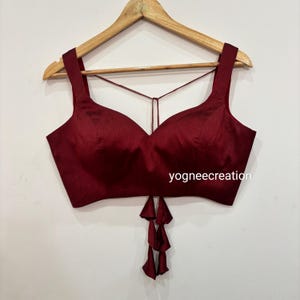 May include: A maroon sleeveless crop top with a deep V-neckline and a tie-up back. The top has a tassel detail at the bottom and the text "yogneecreation" is printed on the front.