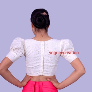 Puede incluir: Blusa blanca de ojal con mangas cortas abullonadas y espalda abotonada. La blusa tiene un dise&ntilde;o perforado delicado y se usa con una prenda rosa. El texto "yogneecreation" es visible.