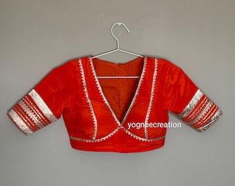 May include: A bright orange blouse with a deep V-neck and short sleeves. The edges and sleeve cuffs are trimmed with silver metallic detailing. The blouse is hanging on a silver hanger against a neutral grey background.